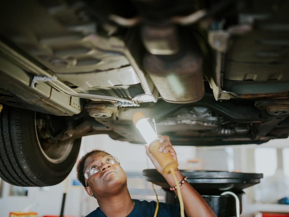 Student working on the underside of a car holding a light
