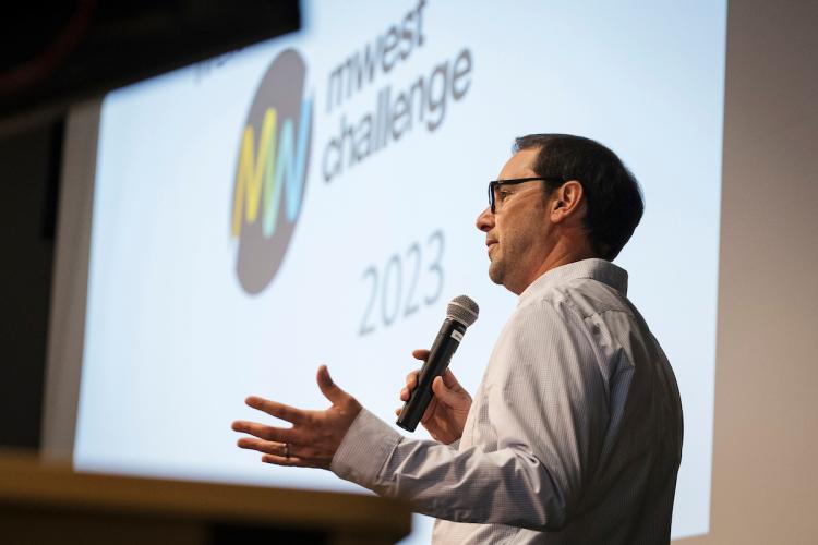 A man stands on stage introducing the MWest Challenge event.
