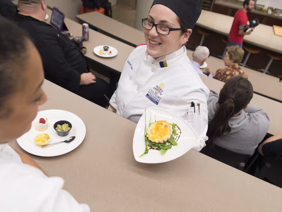 A student chef presenting a dish to a student.