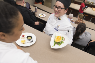 A student chef presenting a dish to a student.