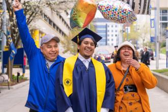 A graduate in a cap and gown poses with two other people, one holding balloons. 