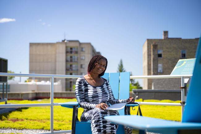 A student sits outdoors on a blue chair with city buildings in the background.