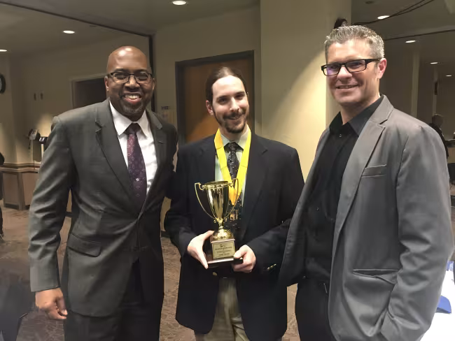 GRCC Andrews Award winner Daniel Cruzan wearing his gold medal and holding his award. GRCC President Bill Pink is on Daniel's right, and professor Andrew Rozema is on his left.