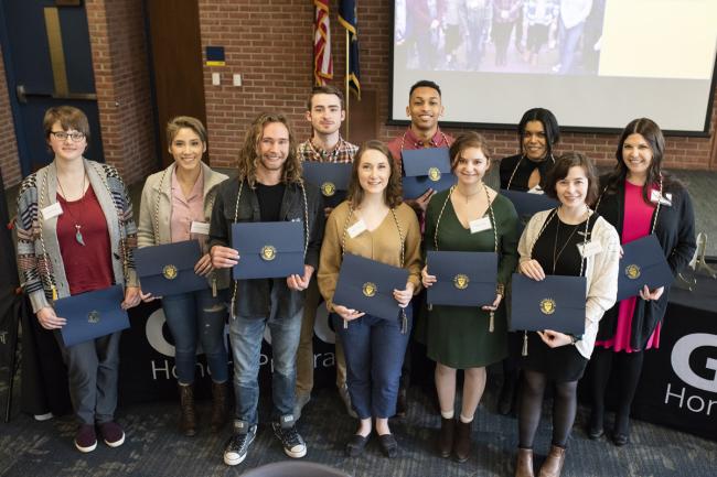 Ten students, all wearing special cords around their necks, hold up their Honors Program certificates as they stand in front of a stage in Sneden Hall.