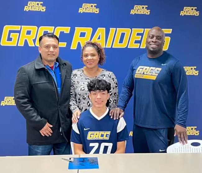 GRCC Jaret Jaramillo sits at a table in front of three people, all are standing in front of a GRCC Raiders banner.