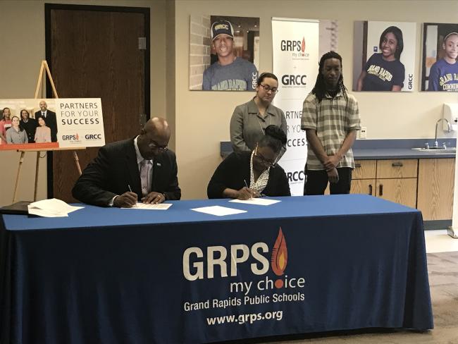 GRCC President Bill Pink and GRPS Superintendent Teresa Weatherall Neal seated at a table signing two documents that exted partnerships for concurrent enrollment and the Learning Center program. Students Najee Smith and Fatemeh Abedi are standing behind them. 