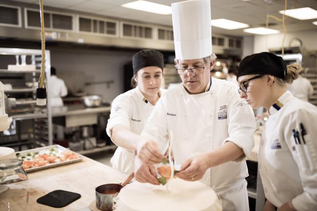Chef Gilles Renusson places a sugar piece on top of a cake as two culinary students watch.