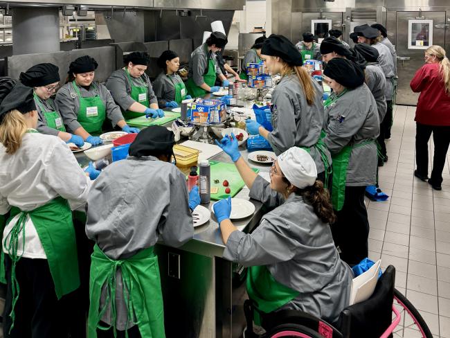 Students and instructors in culinary attire work around a large table in a kitchen.