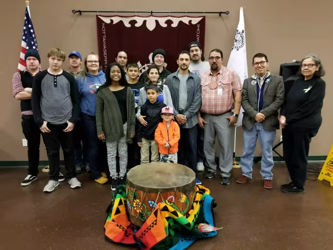 Sixteen people, include two little boys, stand behind a large drum.