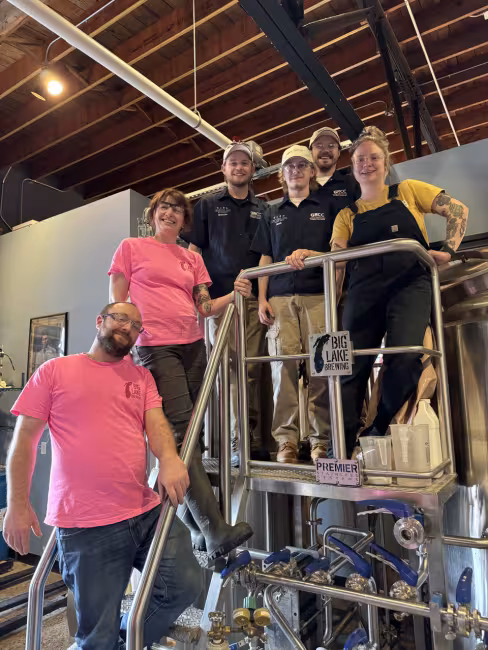 People pose on scaffolding near stainless steel brewing equipment labeled Big Lake Brewing.