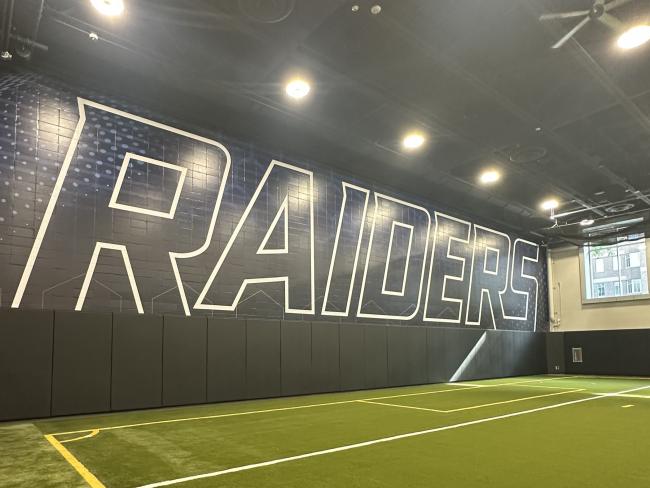An indoor turf field with the word "Raiders" on the wall