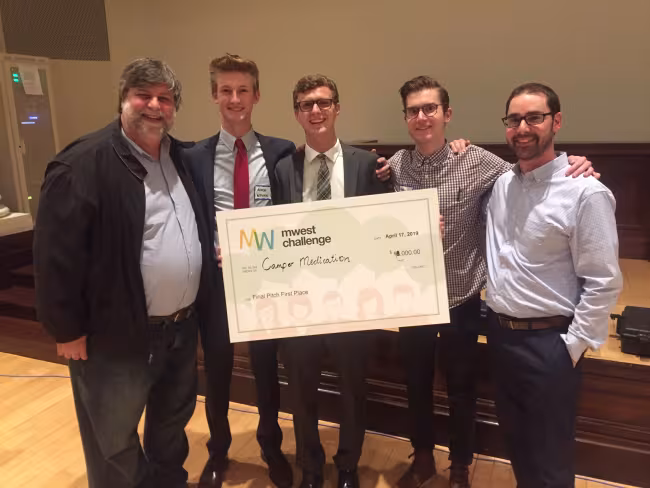 GRCC professor Felix Pereiro stands with ), Aiden Wysocki, Jake Dabkowski, Matthew Veenhoven and Jonathan Moroney, who all hold a giant check.