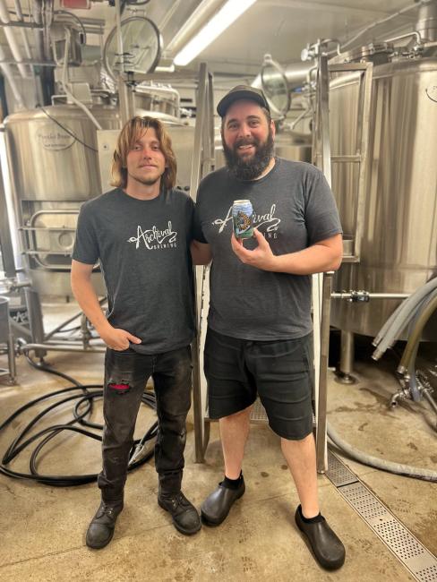 Two people pose in front of steel brewing equipment, holding a beer.
