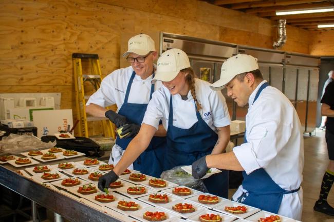 Three people wearing chef's attire work on a large table of food.