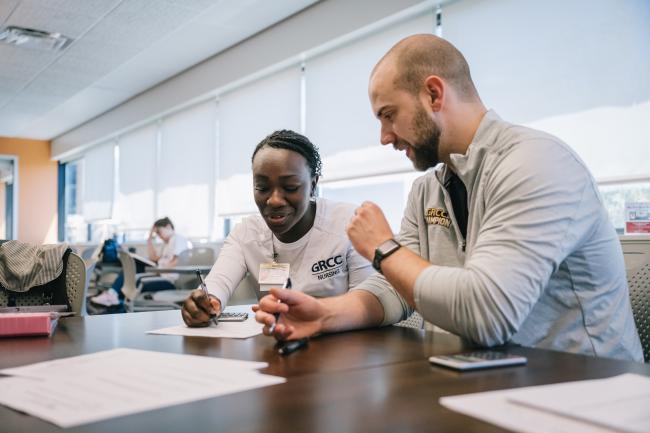A person wearing a GRCC nursing shirt works with another person at a table.