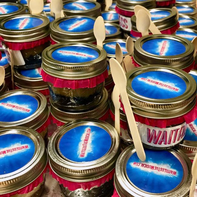 Small pies in a jar with "Waitress" written on the sides and the production website written on the lid. Each glass container has a wooden spoot attached.