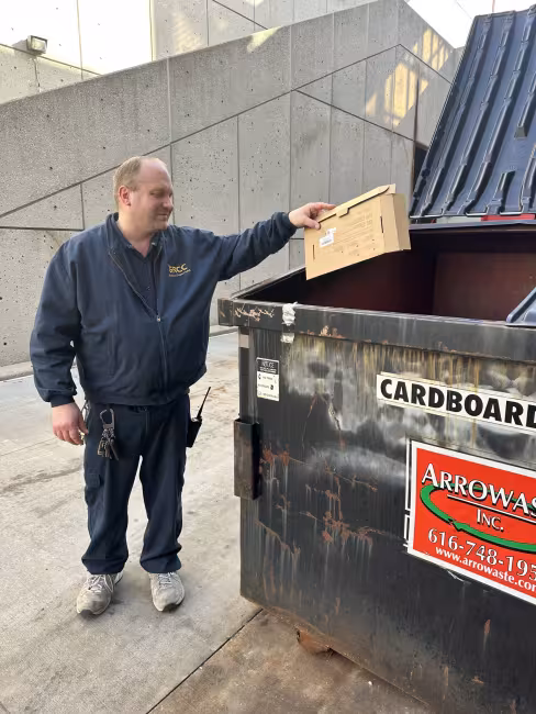 A person drops a cardboard box into a recycling dumpster.