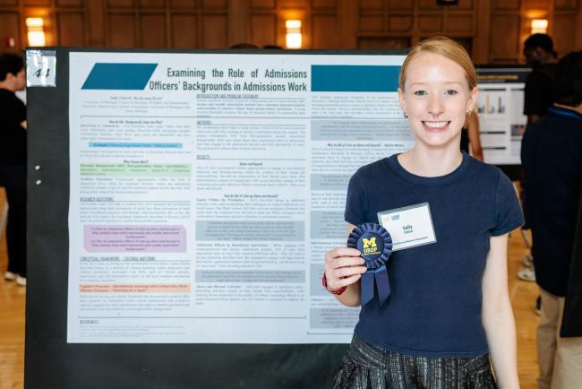 A person stands in front of a poster and holds a blue ribbon.