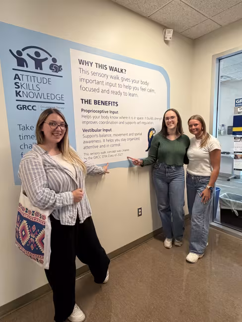 Three students stand outside the OTA department showing off a sign on the wall that explains the sensory walk.