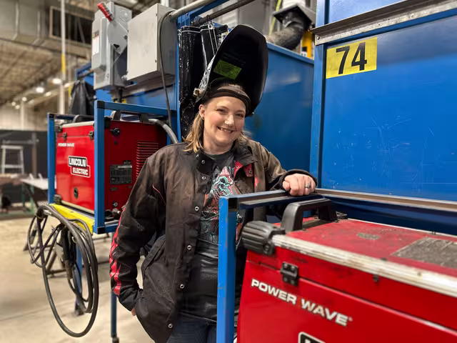 Krista Steffens poses in a welding hat and gear in a manufacturing setting.