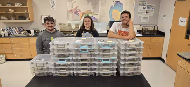 Three students stand behind stacks of plastic containers containing fossils.