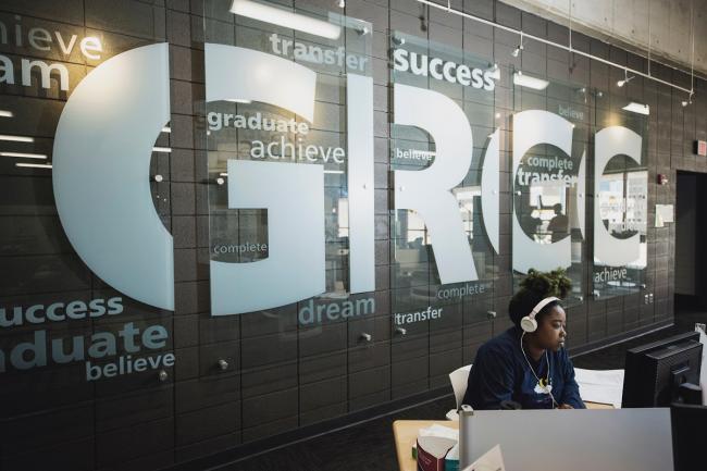 A student sitting in GRCC's academci counseling and advising area, wearing headphones and in front of a wall saying GRCC and encouraging words and phrases.
