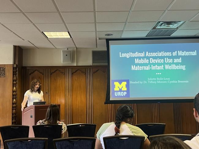 A person presents at a podum in front of a screen reading "Longitudinal Associations of Maternal Mobile Device Use and Maternal-Infant Wellbeing Juliette Bolle-Leon Headed by: Dr. Tiffany Munzer, Cynthia Beauvais"