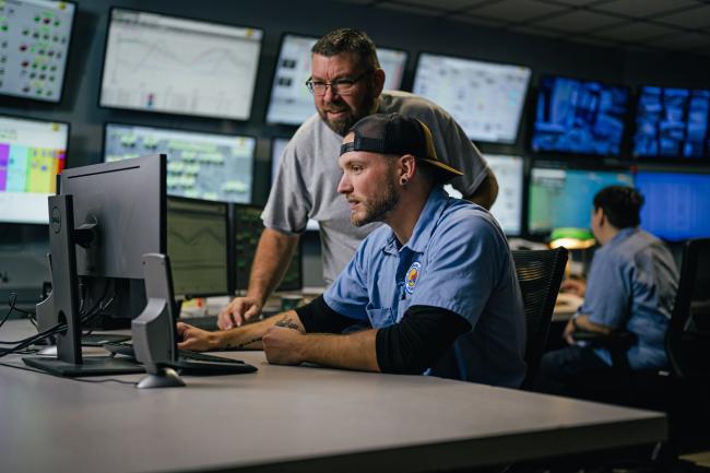 In a room full of monitors, a person looks at a computer screen while somebody looks over his shoulder.