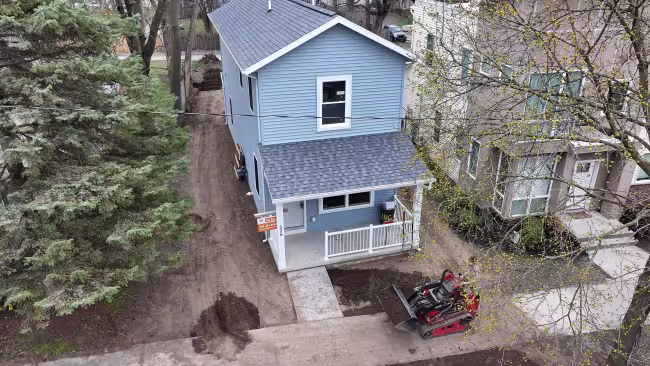 A bulldozer sits outside a two-story, blue house with a porch. 