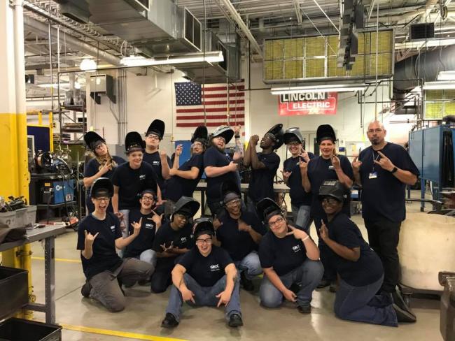 An instructor and 15 welding students, all wearing Metallica Scholars T-shirts and welding helmets, pose in Tassell M-TEC's welding facility.