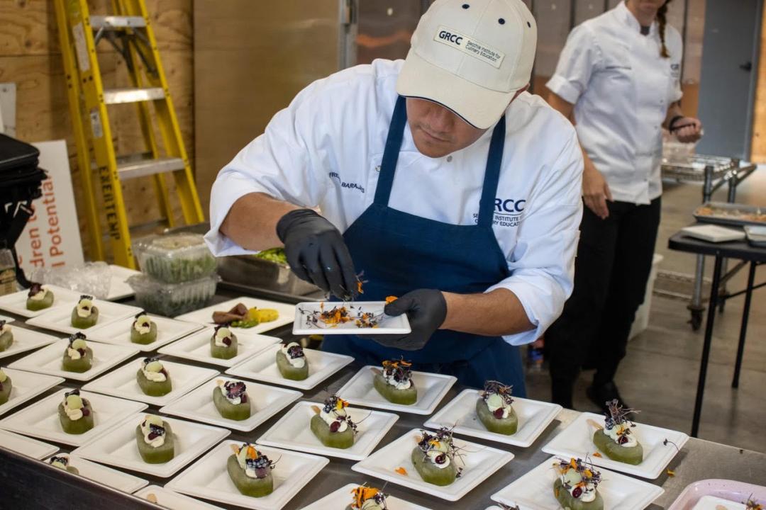 A person wearing GRCC culinary attire prepares many plates of food.