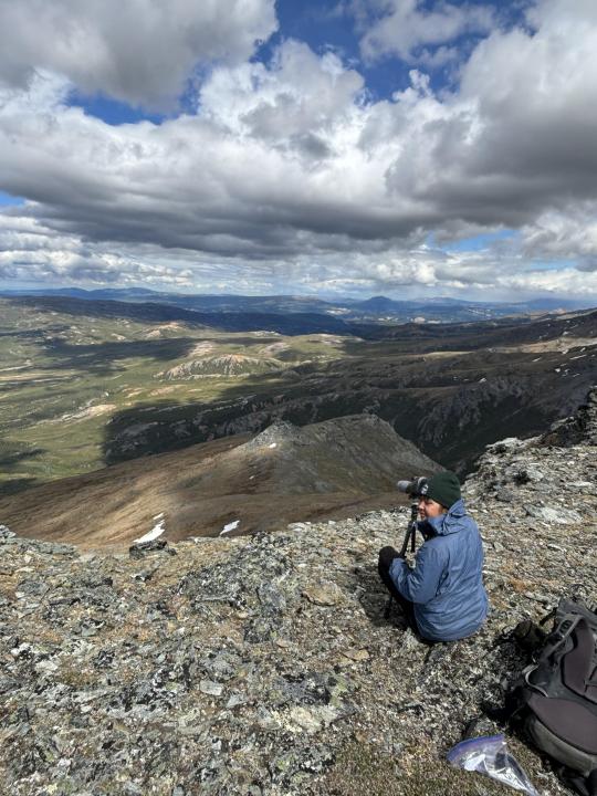 Yasmina sits with a camera on the edge of a mountain overlooking a large valley.