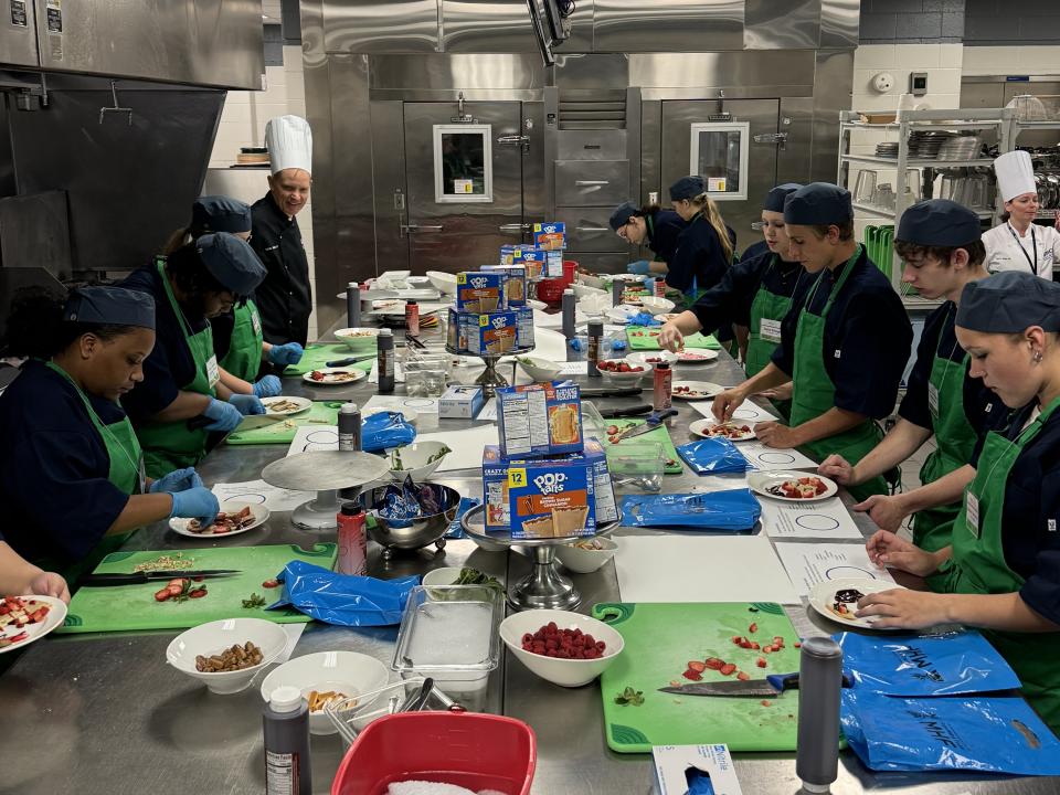 Students and an instructor in culinary attire work around a large table with small plates. In the middle of the table there are ingredients, including a pile of PopTart boxes.