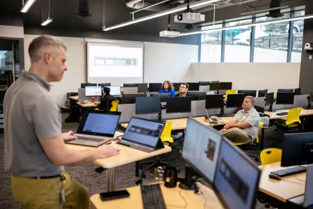 An instructor stands near four computer monitors in a GRCC class. 