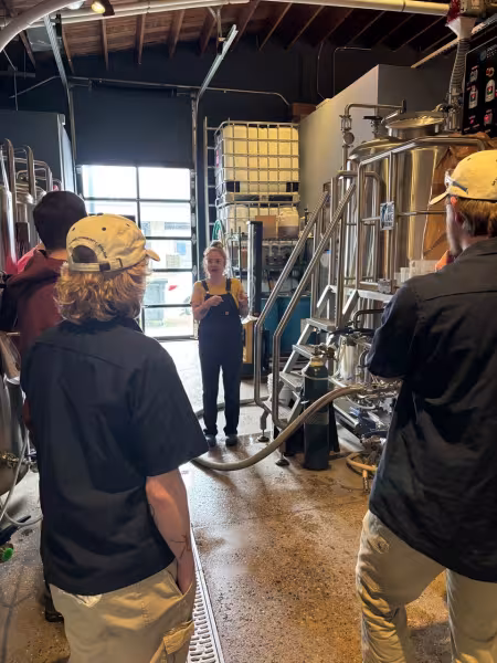 A person speaks to a group of people near brewing equipment.
