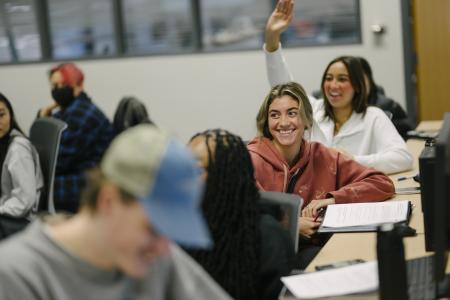 A student in a math class raises her hand while the students around her are smiling.