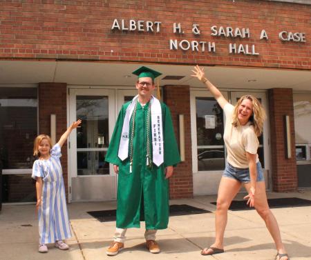 Samuel Wright wears a green graduation gown and cap while his wife and daughter stand on either side, framing him with their arms.