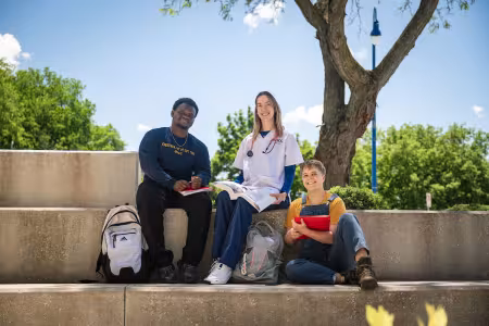Three students sit outside together. One is a nursing student in scrubs.