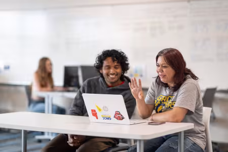 Two students sit in a classroom and work together on a laptop.