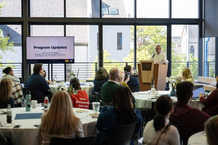 Groups of people seated at tables watch and listen to a presenter talking about the Transfer Bridges program.
