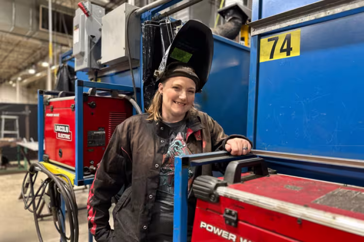 Krista Steffens poses in a welding hat and gear in a manufacturing setting.
