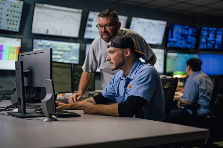 In a room full of monitors, a person looks at a computer screen while somebody looks over his shoulder.