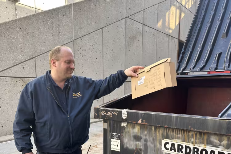 A person drops a cardboard box into a recycling dumpster.