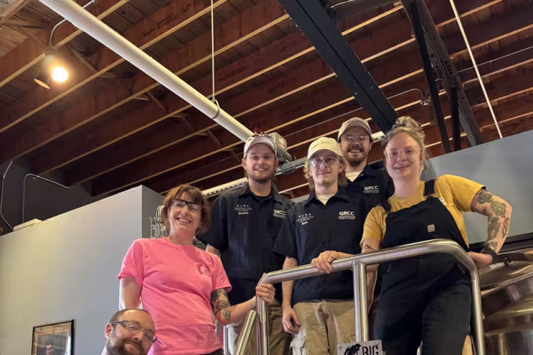 People pose on scaffolding near stainless steel brewing equipment labeled Big Lake Brewing.