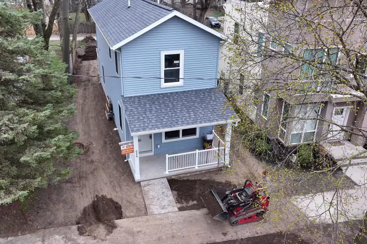 A bulldozer sits outside a two-story, blue house with a porch. 