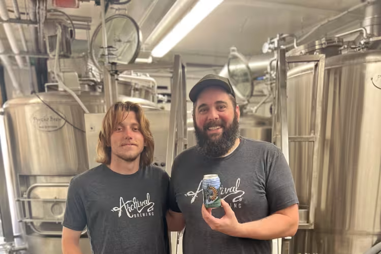 Two people pose in front of steel brewing equipment, holding a beer can.
