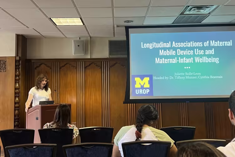 A person presents at a podum in front of a screen reading "Longitudinal Associations of Maternal Mobile Device Use and Maternal-Infant Wellbeing Juliette Bolle-Leon Headed by: Dr. Tiffany Munzer, Cynthia Beauvais"