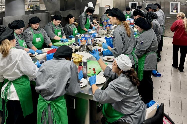 Students and instructors in culinary attire work around a large table in a kitchen.