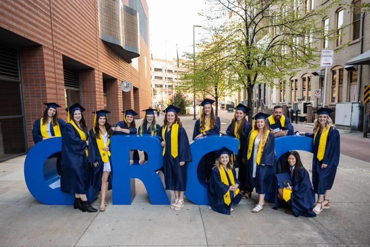 A group of graduates in caps and gowns pose around big blue letters spelling out GRCC.