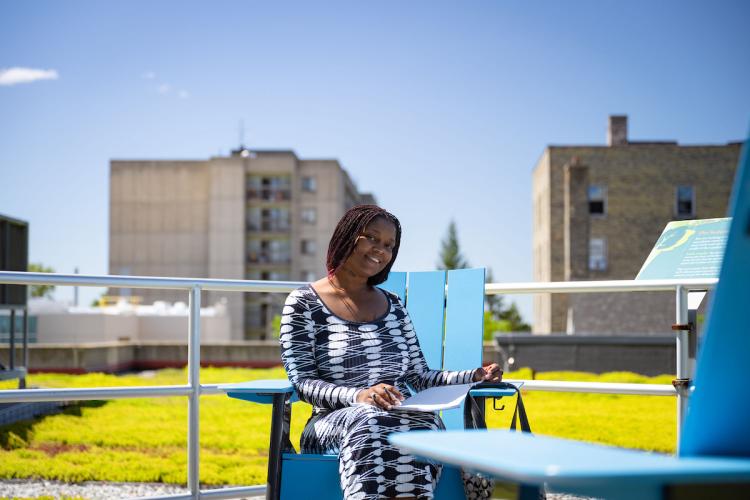 A student sits outdoors on a blue chair with city buildings in the background.
